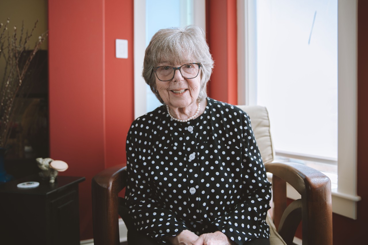 Margaret Oulton is seated indoors in a black blouse with white polka dots and smiling.