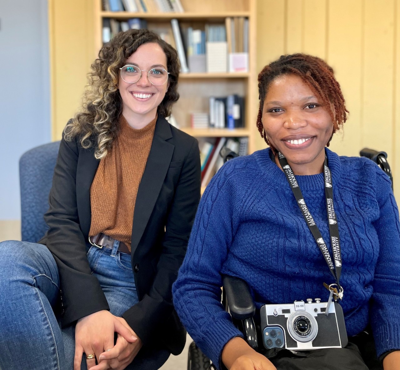 Two women are seated, facing the camera straight on, and smiling. The woman on the left is white, has curly brown hair and glasses, and is wearing a black blazer over a caramel-coloured shirt and blue jeans. The woman on the right is Black, and seated in a motorized wheelchair. She has short, kinky hair and is wearing a blue sweater with a Dalhousie lanyard.