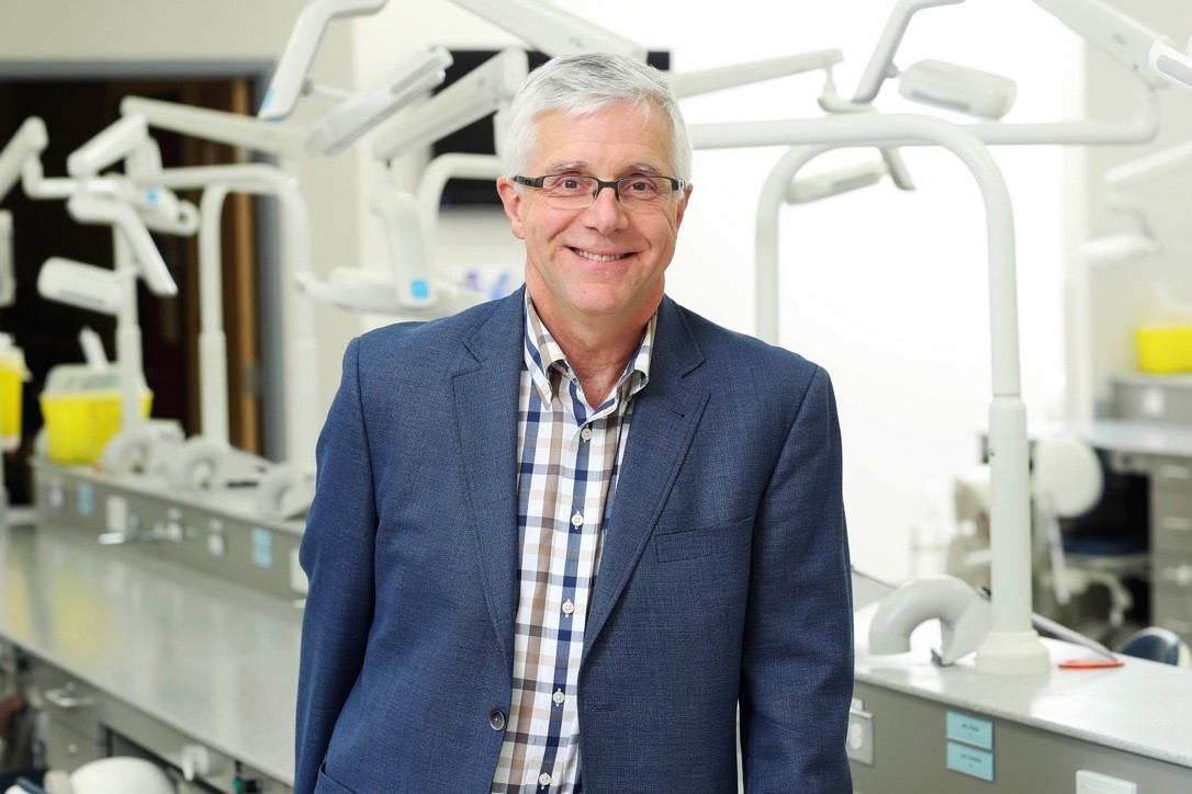Dr. Lee Erickson stands smiling in a dental lab dressed in business casual attire.