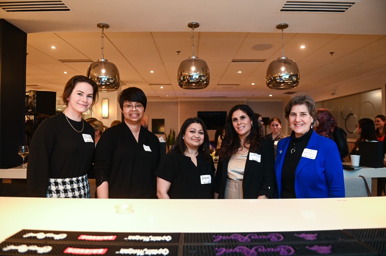 Five female-presenting women dressed in business casual attire wear nametags at an event in a restaurant setting.
