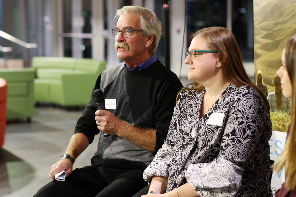 Swindon is seated and speaks into a microphone next to a woman in a large room with light green couches. 