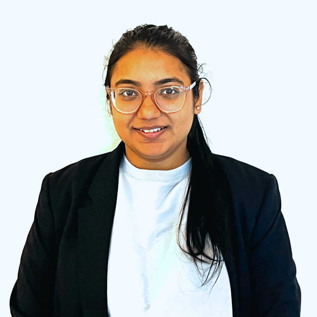 An Indian woman faces the camera straight on. Her long dark hair is pulled into a ponytail that hangs over one shoulder. She is wearing large, clear-framed glasses and a black blazer with a white T-shirt underneath. She is smiling.