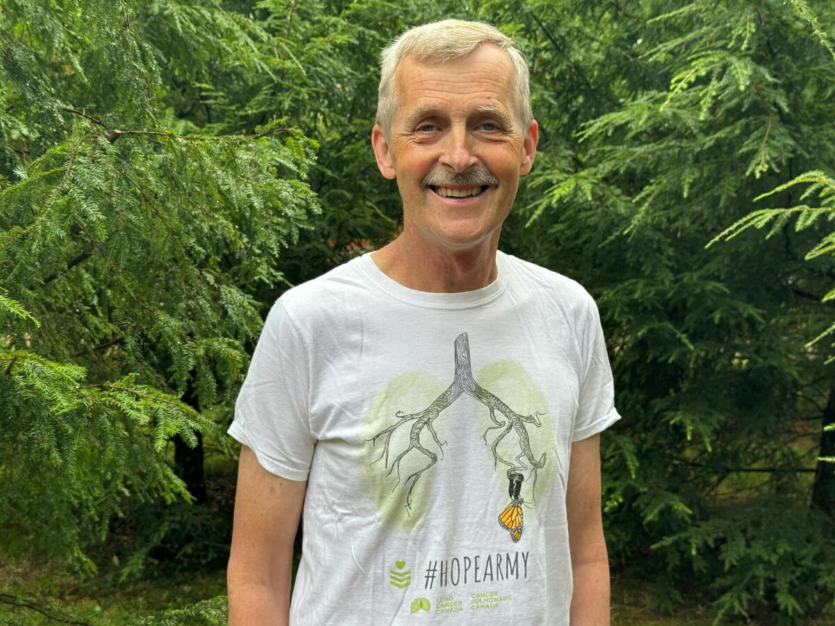 A man dressed casually smiles outdoors in fine weather against a backdrop of trees.