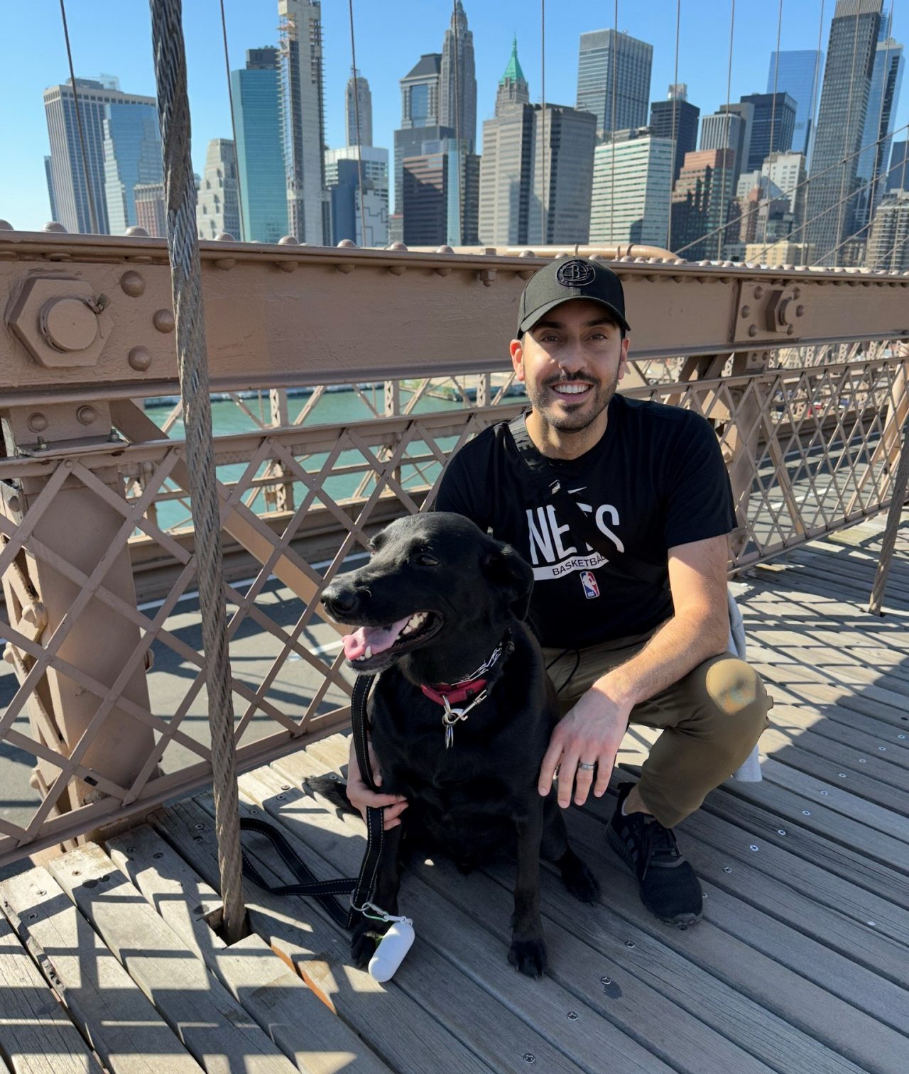 Ahluwalia is crouched on the bridge smiling with a black dog and the Brooklyn skyline in the background.
