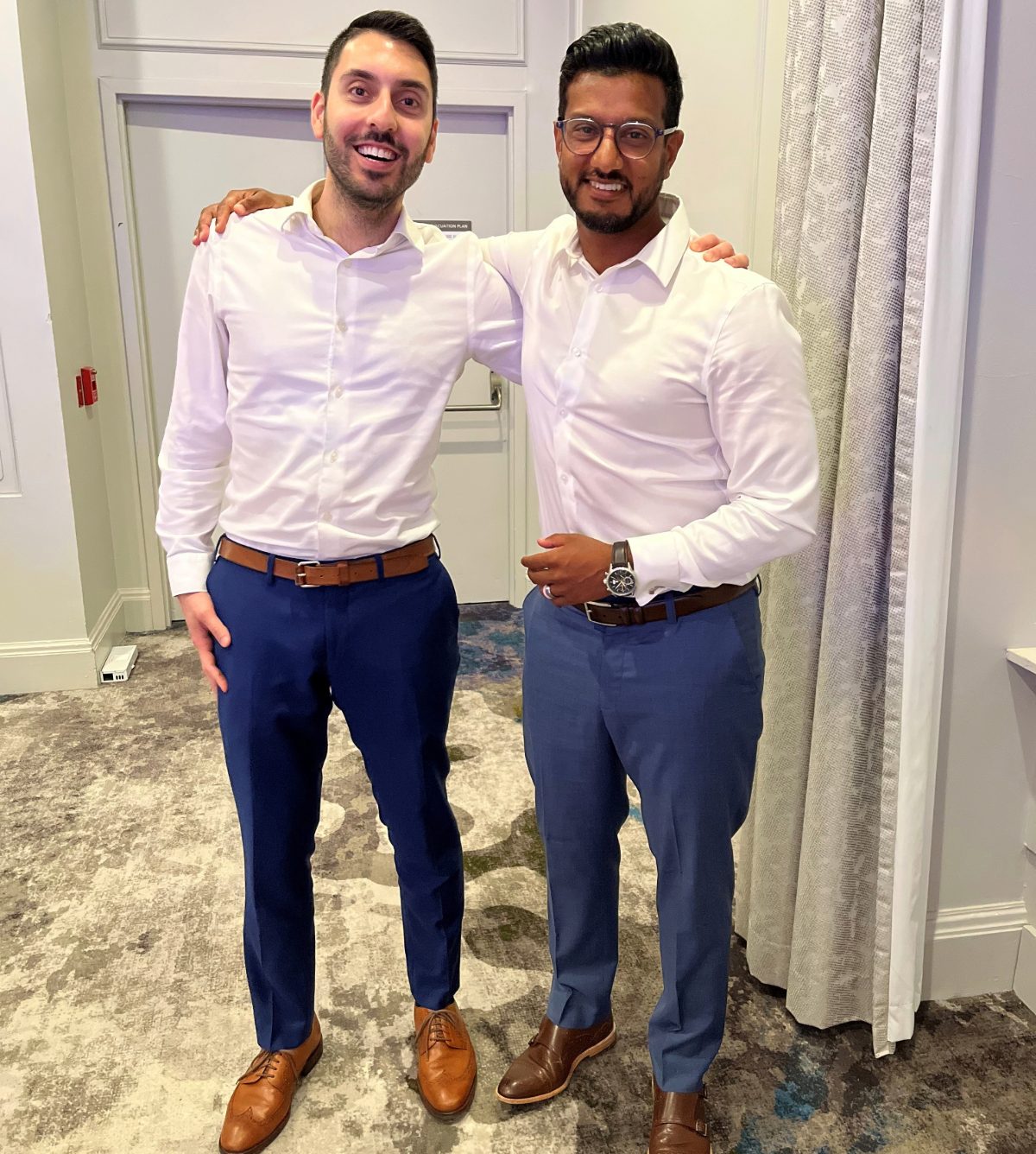 Two men dressed semi-formally in white shirts, blue pants and brown shoes stand indoors with their arms around each other's shoulders.