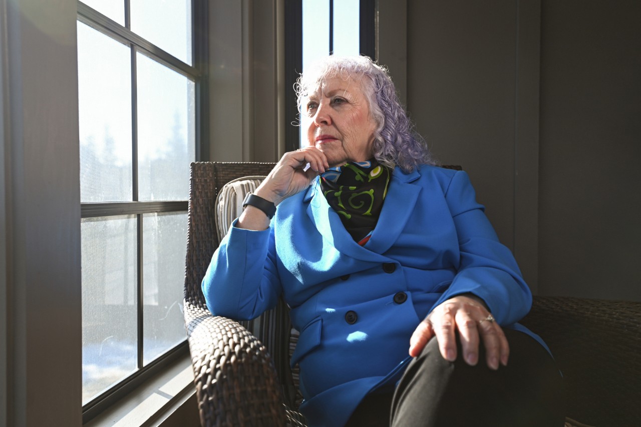 Lynch is seated looking out a window with her hand at her chin and wearing a blue jacket and colourful scarf.