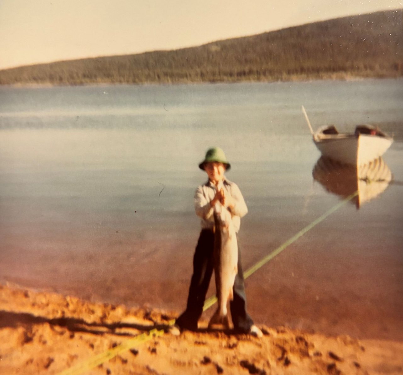 An archival photo of a child holding up a large fish standing on the shore of a lake.