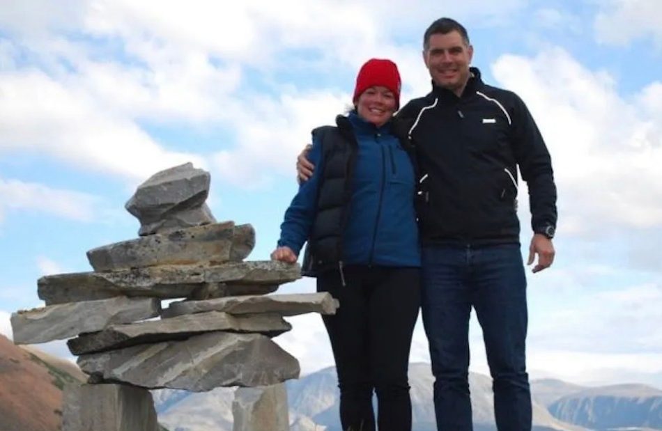 Two people stand beside a small stack of stones on a mountaintop with the landscape and cloudy sky in the background.