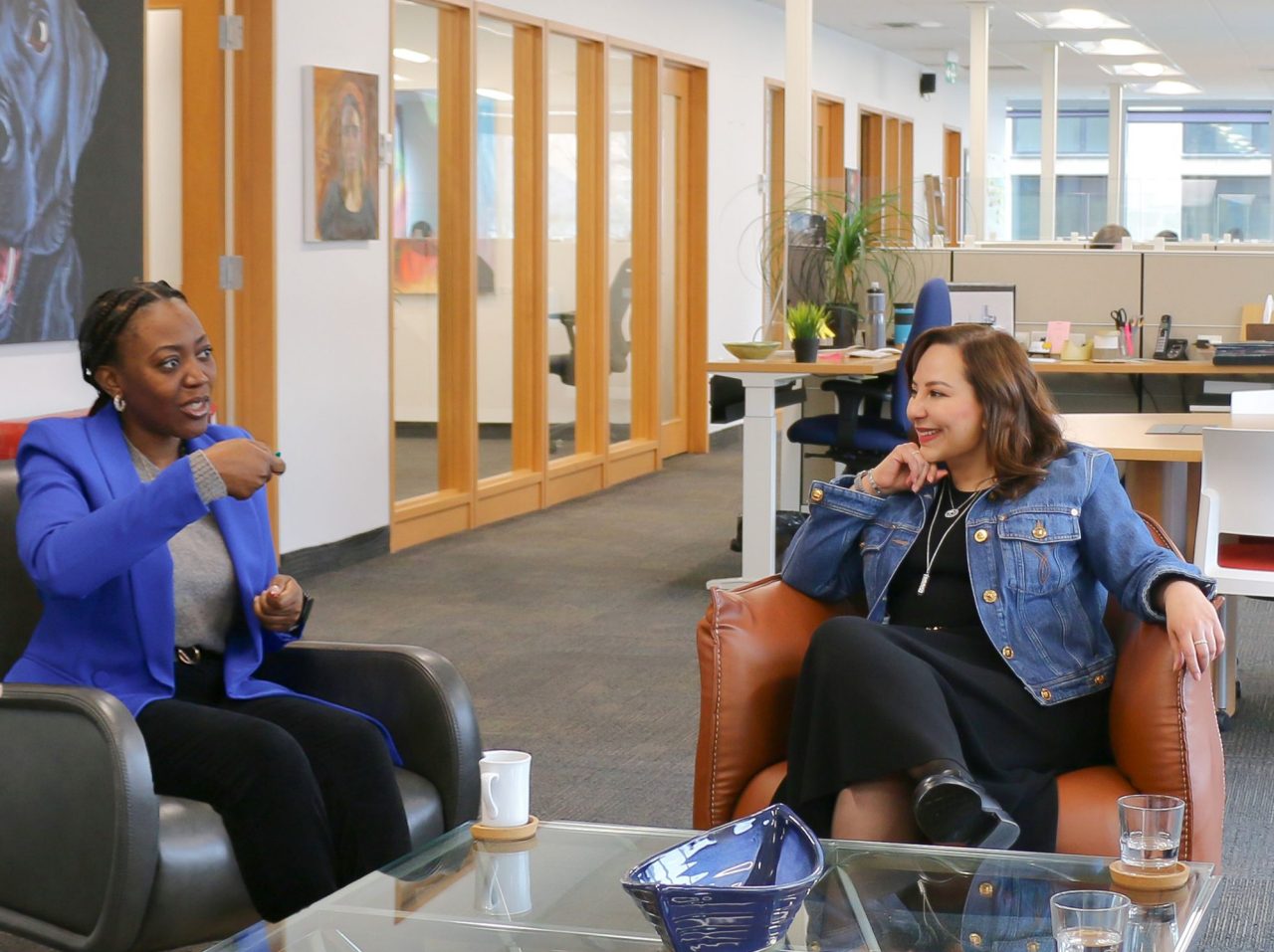 Two women sit in conversation in an open office space. 