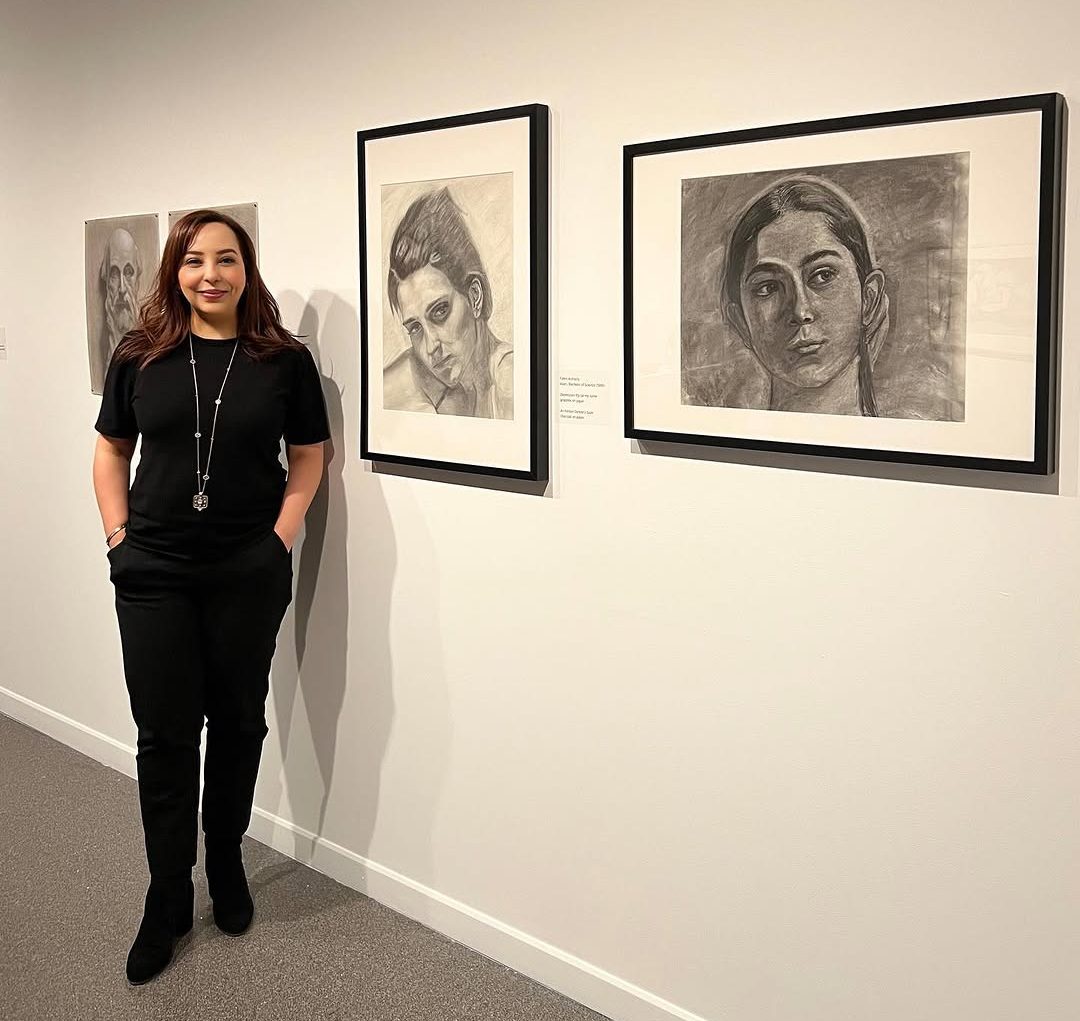 Faten Alshazly standing next to several framed artworks displayed on a wall.