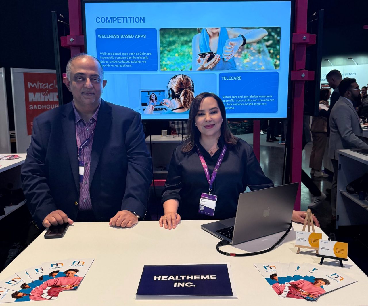 Two people sit at a booth in front of a large screen at a conference tradeshow.