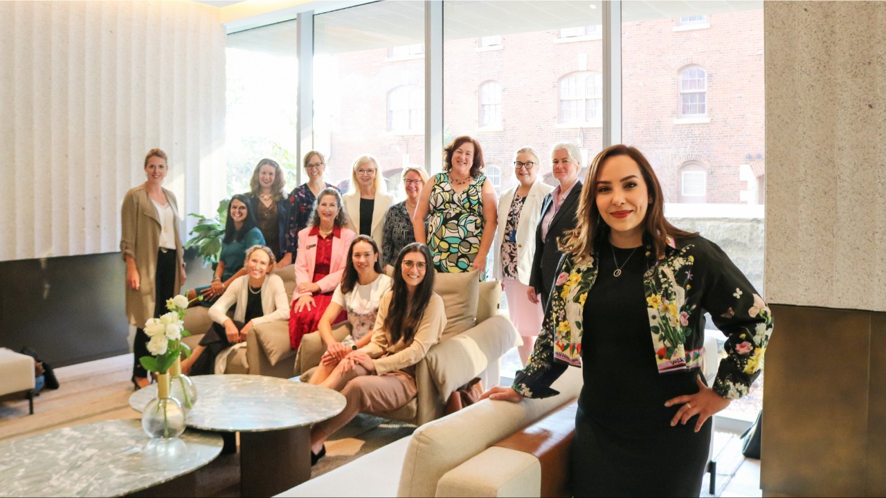 A woman stands smiling in a bright lounge space with a group gathered in the background.