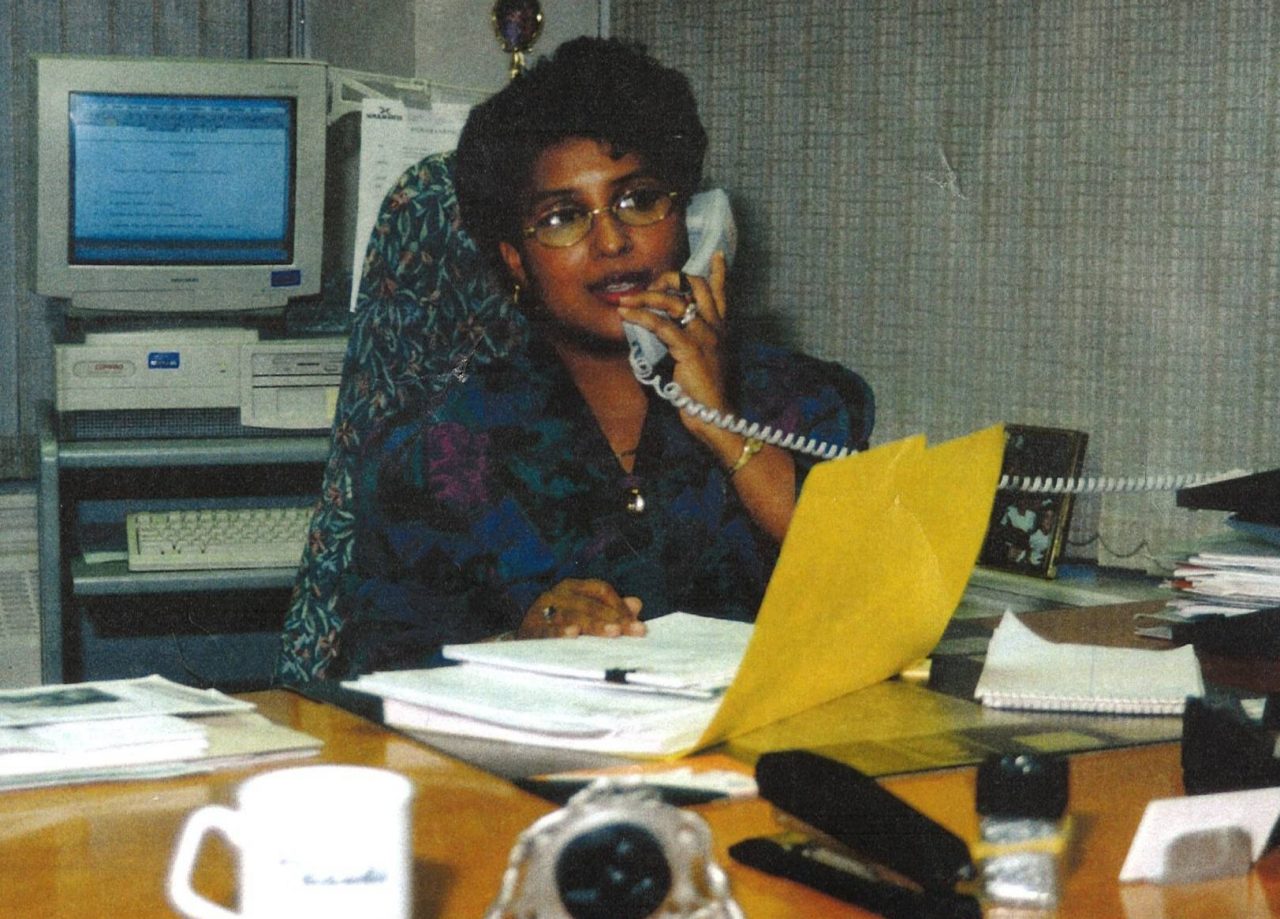 A woman talking on a corded phone sitting at a desk with a desktop computer circa 1980s. 