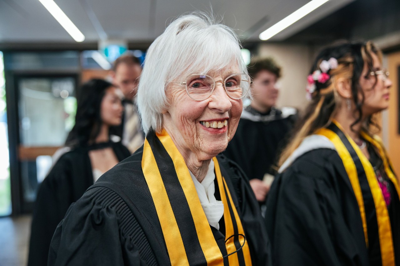 A mature woman smiling widely in convocation gown amongst other graduands nearby.