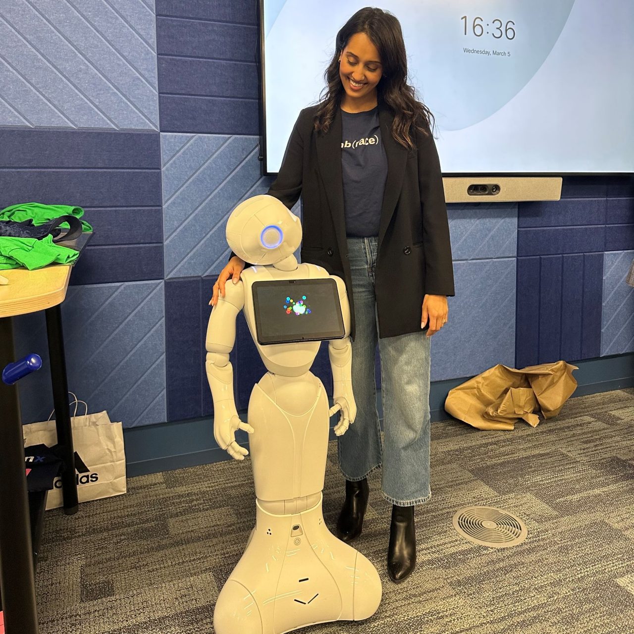 A woman stands next to a robot about waist-high in a somewhat human form with her hand on its shoulder as they appear to look at each other in an indoor setting.