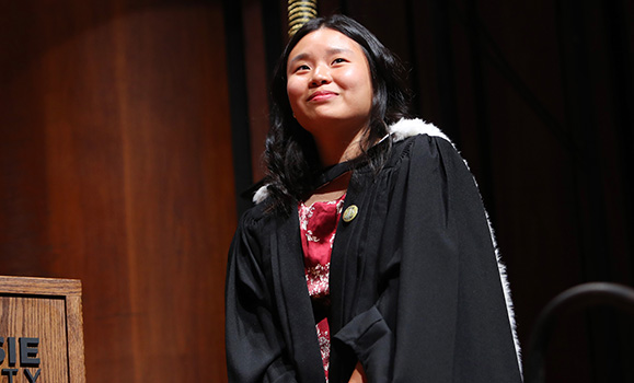A woman in graduation gown smiles slightly from the stage.