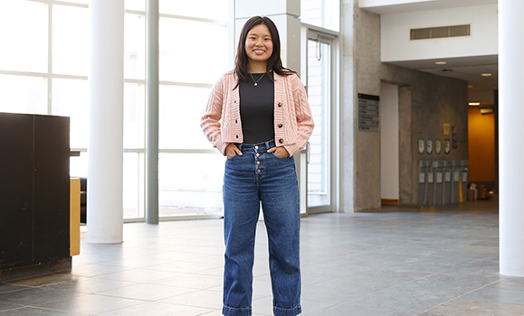 Michelle Wang stands smiling with her hands loosely in her jeans pockets indoors in a well lit indoor space.