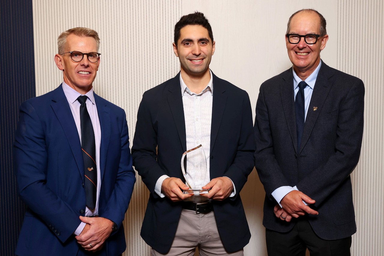 A man stands holding a crystal award smiling between two other men at an event.. 