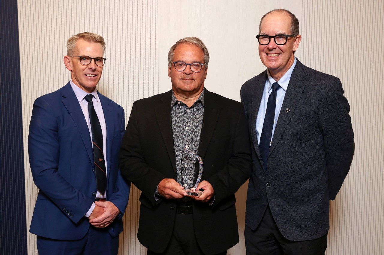 A man stands holding a crystal award smiling between two other men. 