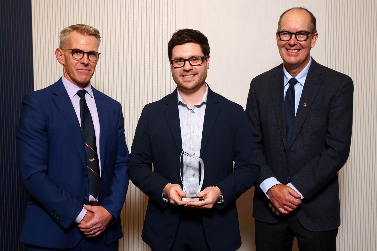 A man stands holding a crystal award smiling between two other men. 