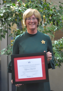 Karen Oldfield stands holding a framed certificate at an event.
