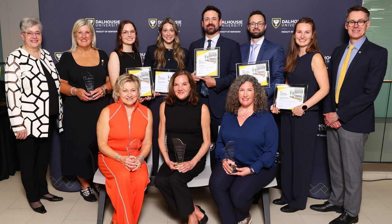 Award recipients stand holding framed certificates or crystal awards alongside several others standing and seated.