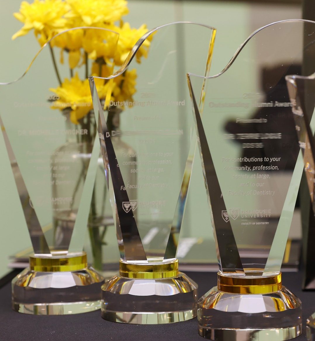 Four crystal awards etched with recipient information lined up on a table with a vase of yellow flowers.