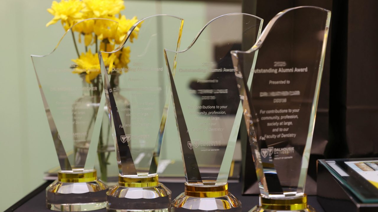 Four crystal awards etched with recipient information lined up on a table with a vase of yellow flowers.