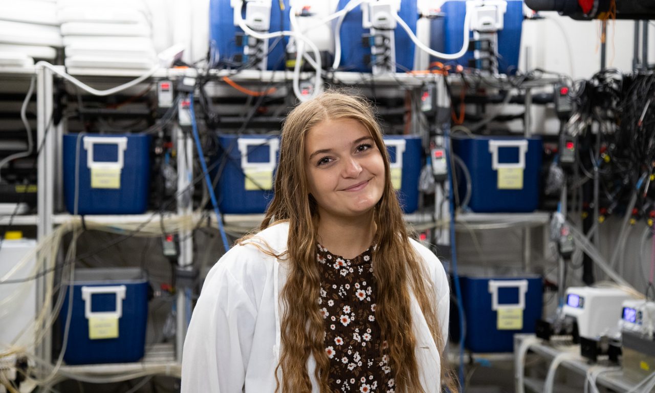 A female student with long hair stands smiling surrounded by equipment on shelves in a lab.