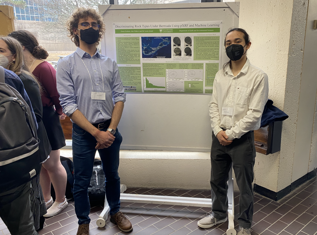 Two male students wearing masks stand with their hands clasped in front of them with a presentation board behind them in an indoor space. 