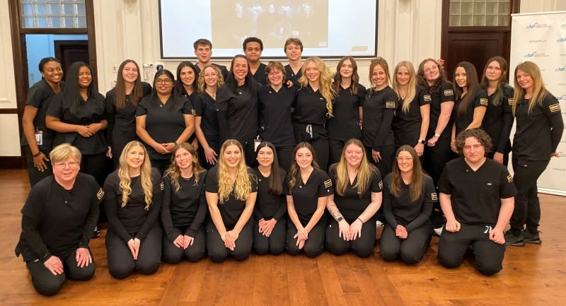 A large group of nursing students posed together at an event in a large hall with a screen behind them showing a photo of  a previous class.