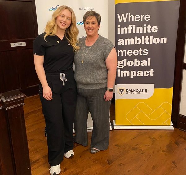 A daughter and mother stand smiling together at an indoor event with banners behind them.