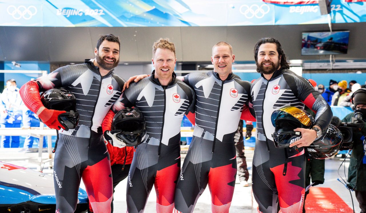 Four men wearing bobsleigh race suits and holding helmets stand with their arms around each other smiling in an arena.