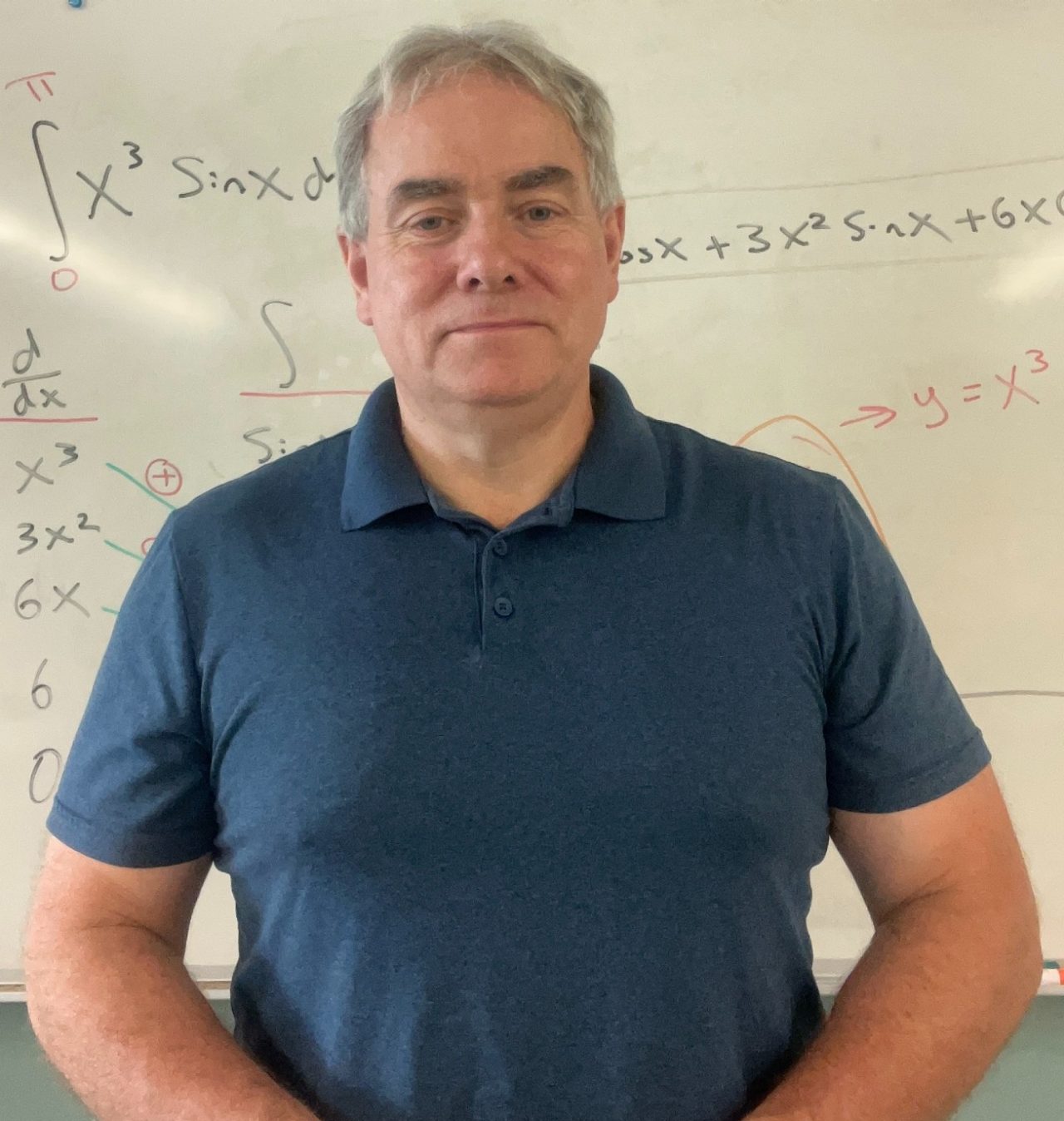 Frank Brown standing in a classroom smiling slightly in front of whiteboard.