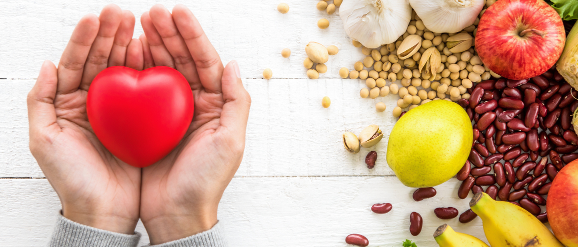 A red heart object lies in the palms of two hands with a collection of fresh food in the background.