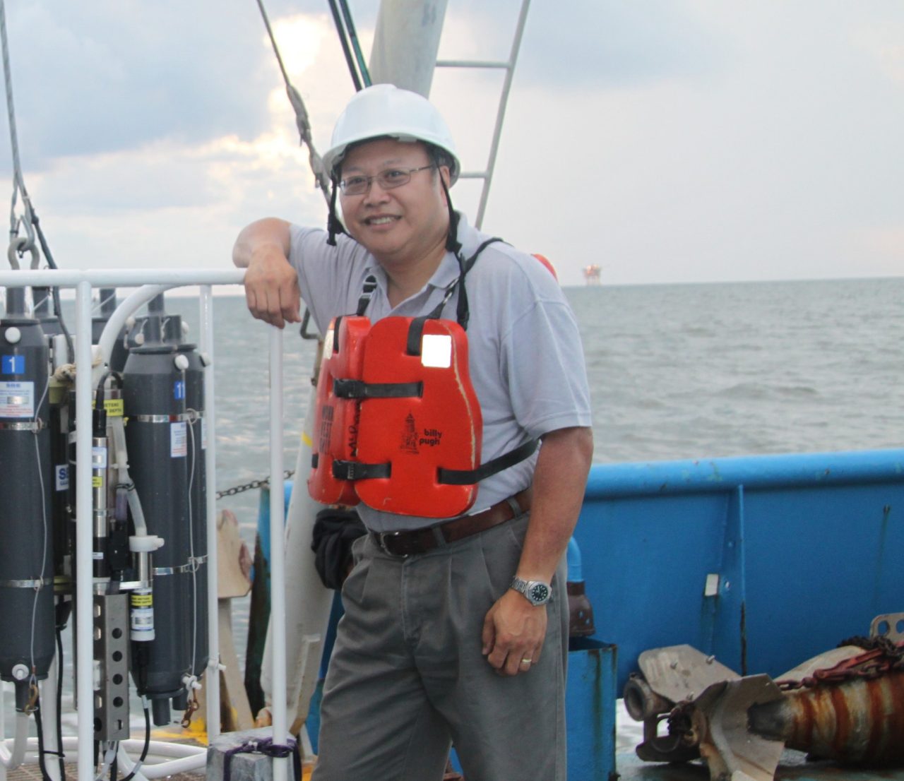 A man stands smiling aboard a ship at sea wearing a life preserver and a hard hat.