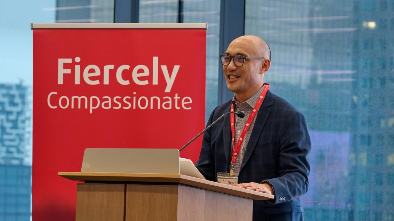 Person speaking at a podium beside a red “Fiercely Compassionate” sign with large windows and a cityscape in the background.