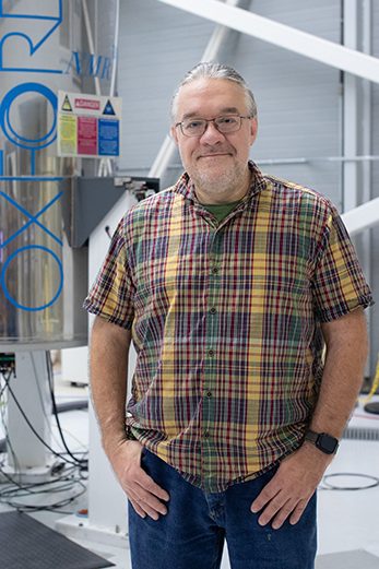 Person standing in a laboratory beside a large stainless steel tank labeled “OXFORD,” with equipment, cables and workbench in background.