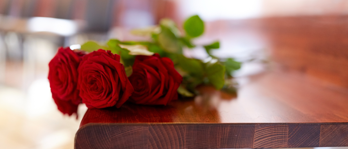 Two red roses sit on a wooden table in front of a row of chairs.