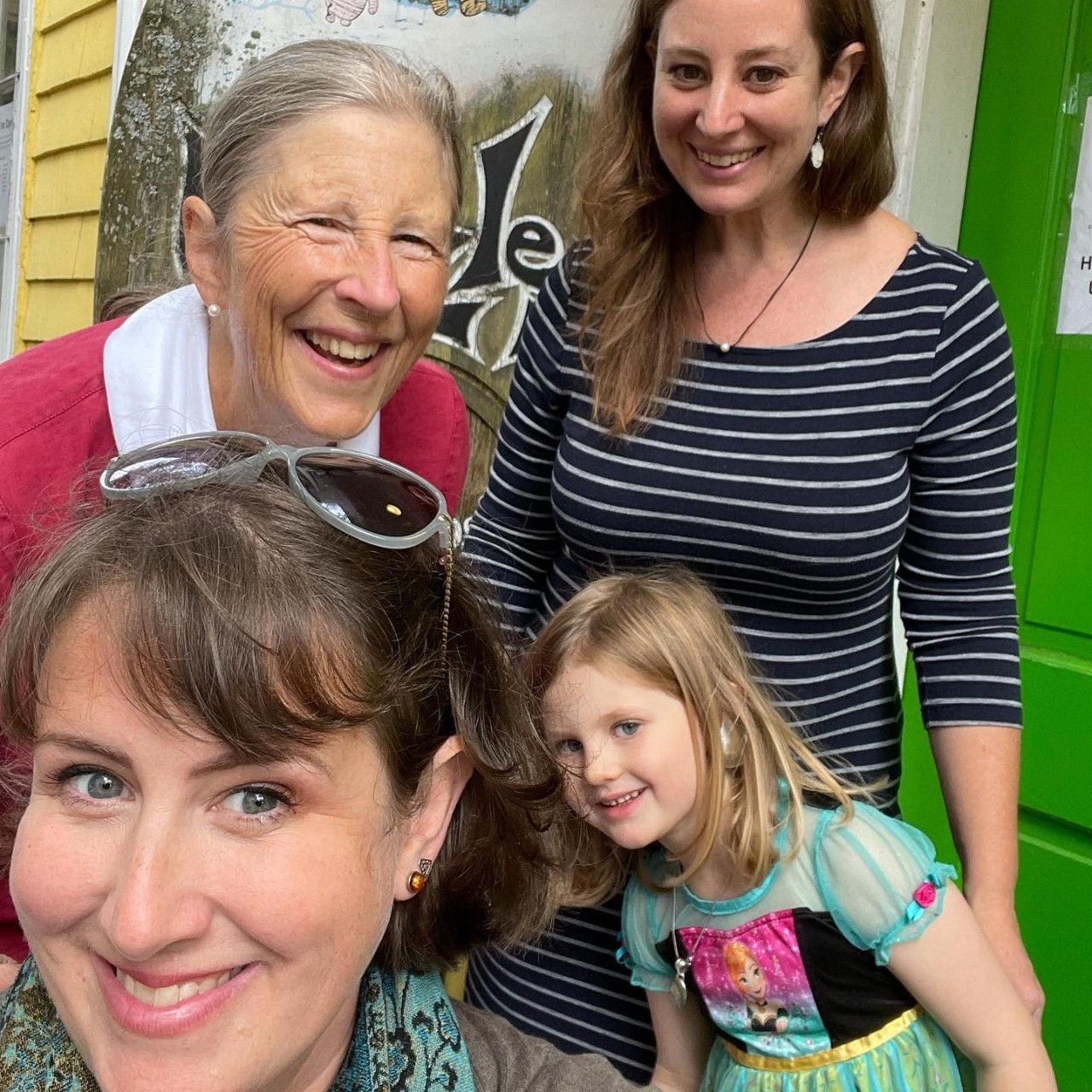 Four women from three generations smile together on the doorstep to a store.