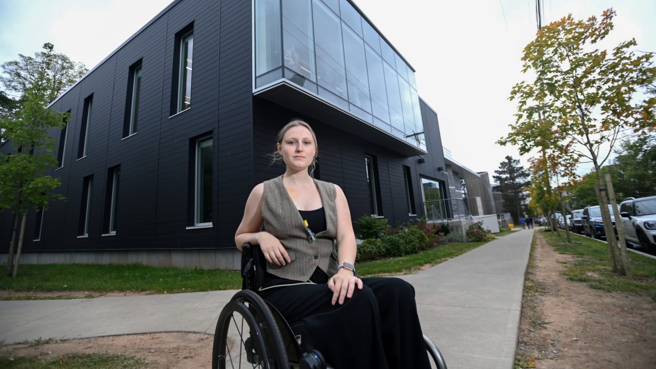 Taverner sits in a wheelchair on the sidewalk outside the Strug Concert Hall on a tree-lined street.