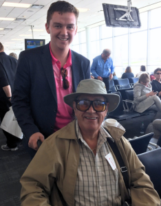 Aaron Prosper stands behind Elder Albert Marshall who is seated in an airport waiting area setting. 
