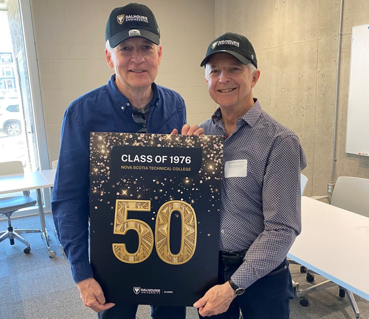 Two members of the engineering class of 1976 hold a sign with a gold "50" and wear Dalhousie ball caps.