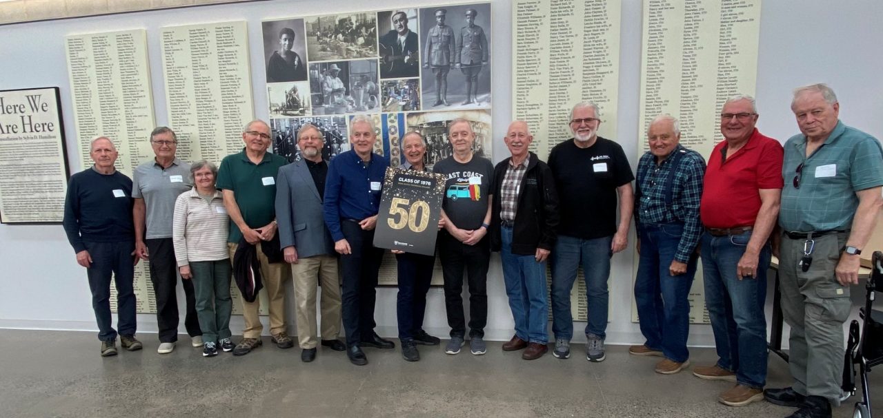 A group of people standing in a row with one holding a gold "50" sign and historic photos and lists of names on displays behind them.