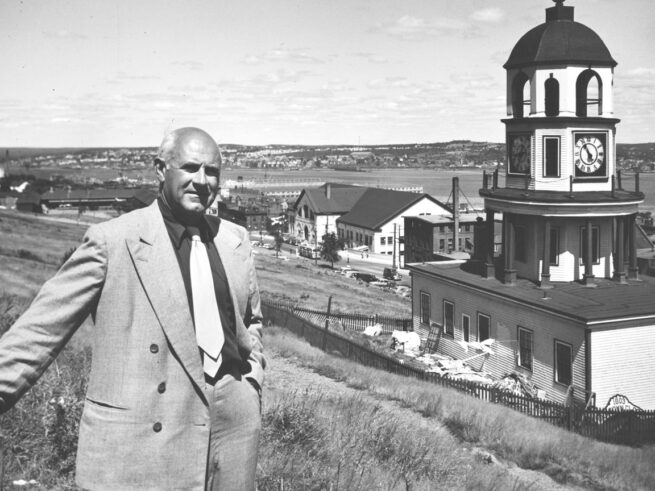 An archival black-and-white photograph of a balding man in a suit standing on Citadel Hill near the clocktower. The harbour and some small buildings are visible behind him.