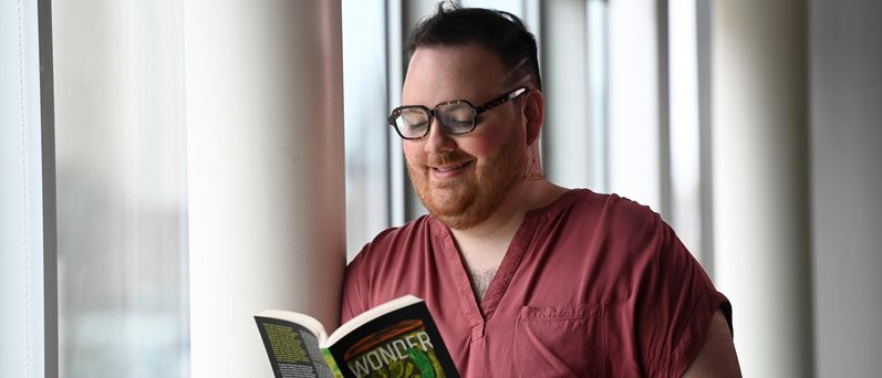 A non-binary person with a beard and black glasses is wearing a red shirt and leaning against a white post. They are smiling and looking at the open book in their hands, which is black and green and titled "Wonder World."