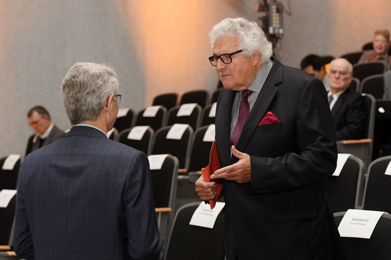 Two men dressed in business formal attire in conversation in an auditorium with several other people seated around them.