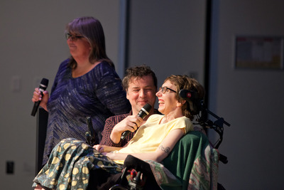 A thin white woman sits reclined in a motorized wheelchair, smiling broadly at something off camera. A white man kneels beside her, smiling warmly, and holds a black microphone in front of her. A woman with grey hair and a second microphone stands in the background.