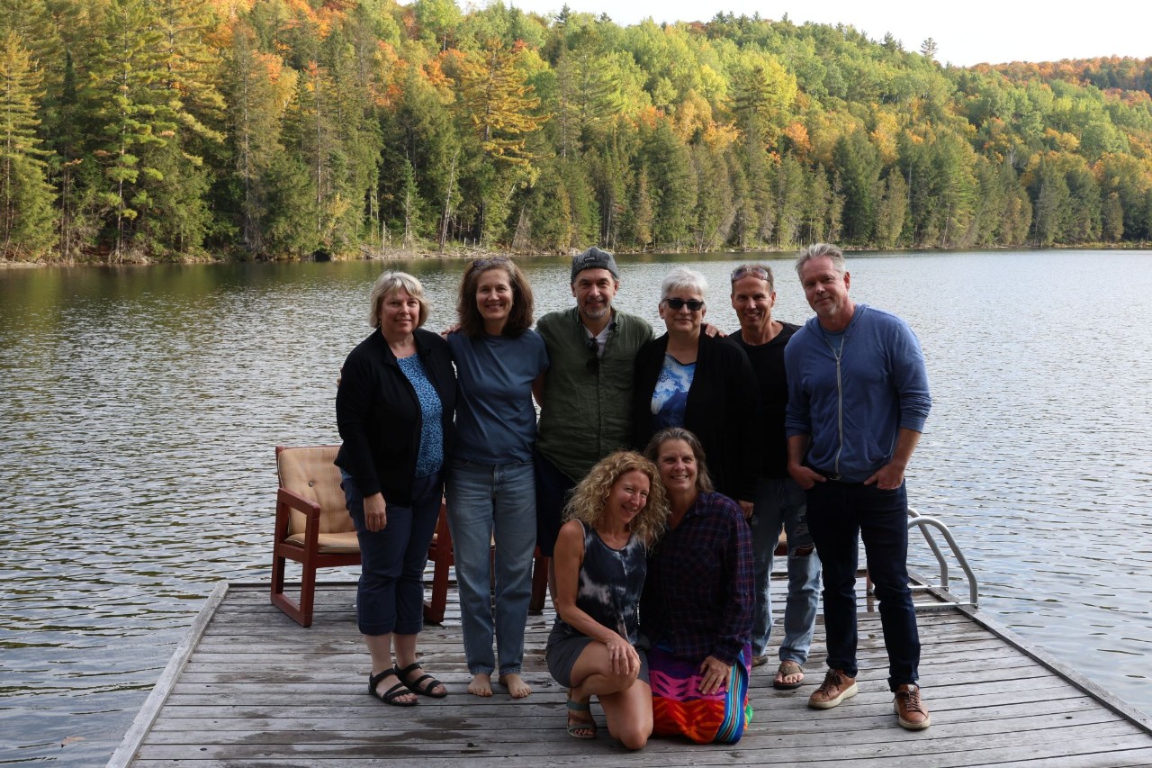 A group of eight men and women dressed casually in early fall are gathered together at the end of a dock over water with a background of trees.