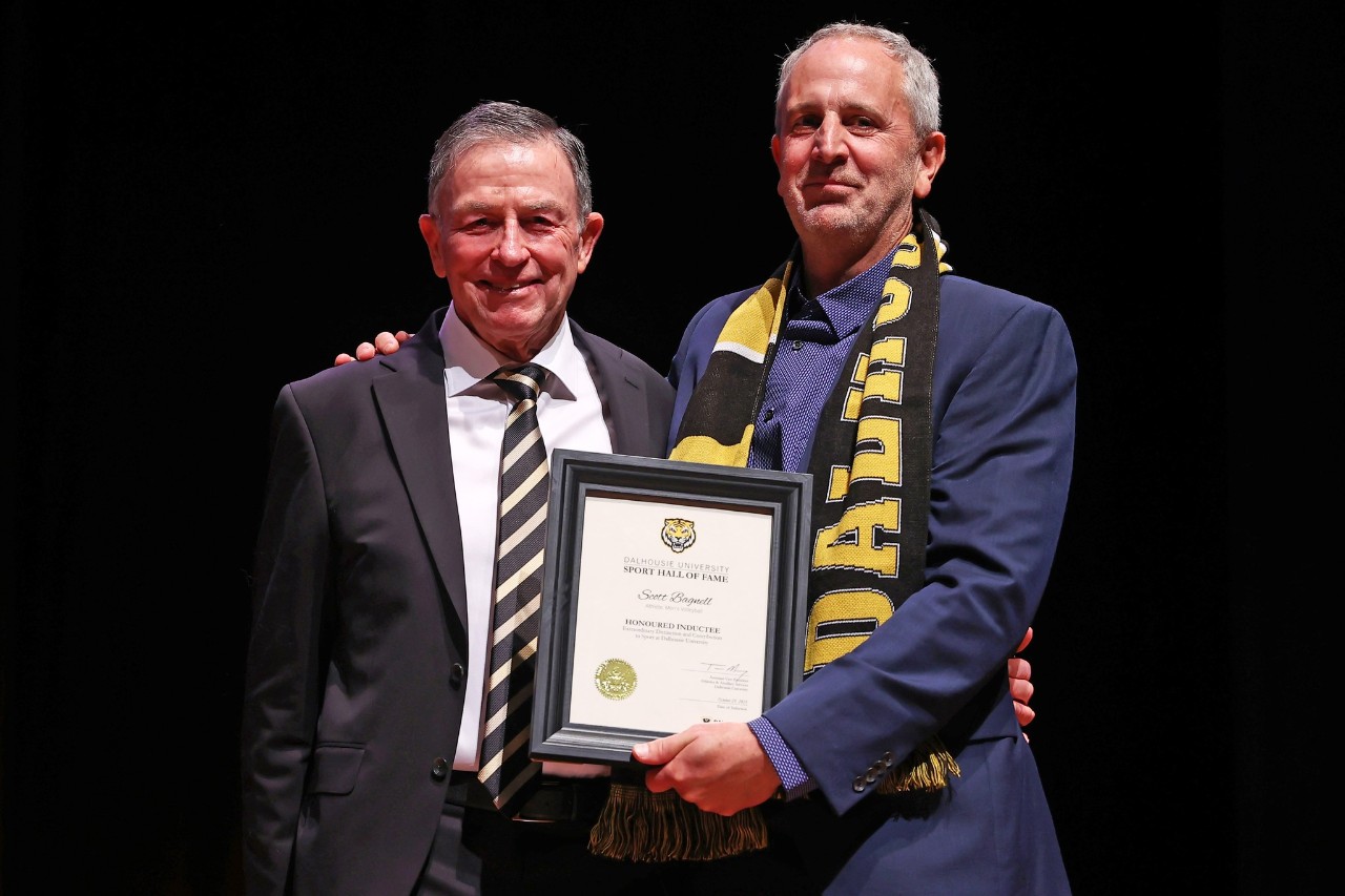 A man wearing a black and gold scarf holds a framed certificate standing next to a man in a suit smiling on stage at an event.